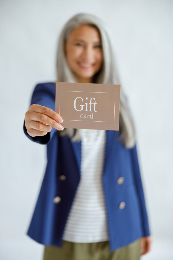 Positive middle aged Asian woman on light grey background, focus on hand with gift card